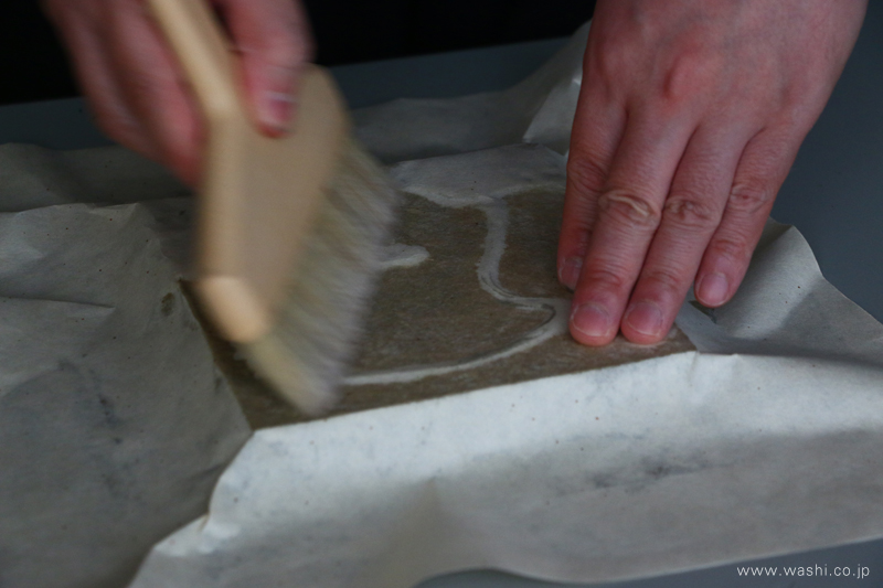 和紙アートパネルの制作風景。職人が刷毛を使い、パネルに和紙を丁寧に貼り込んでいる手仕事の様子。