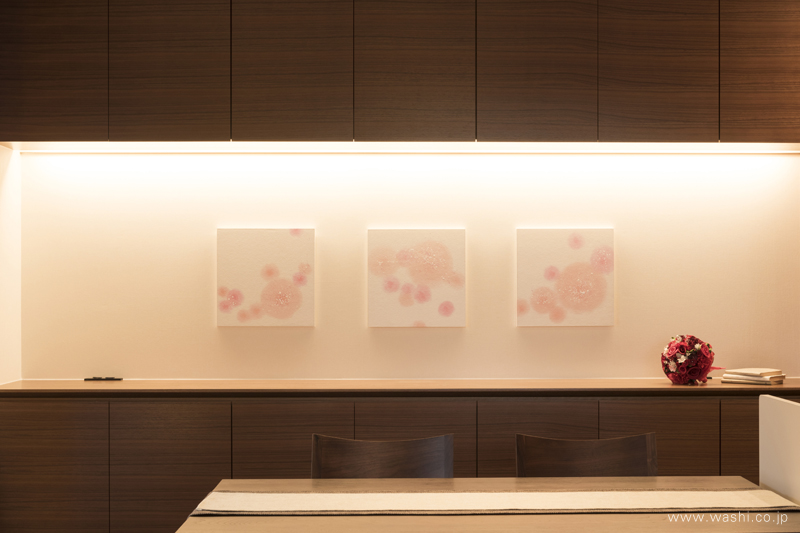 木目調のダイニングに飾られた、桜を思わせるピンク色の丸い模様の和紙アートパネル3枚。間接照明に照らされ、空間を明るく華やかに彩っています。